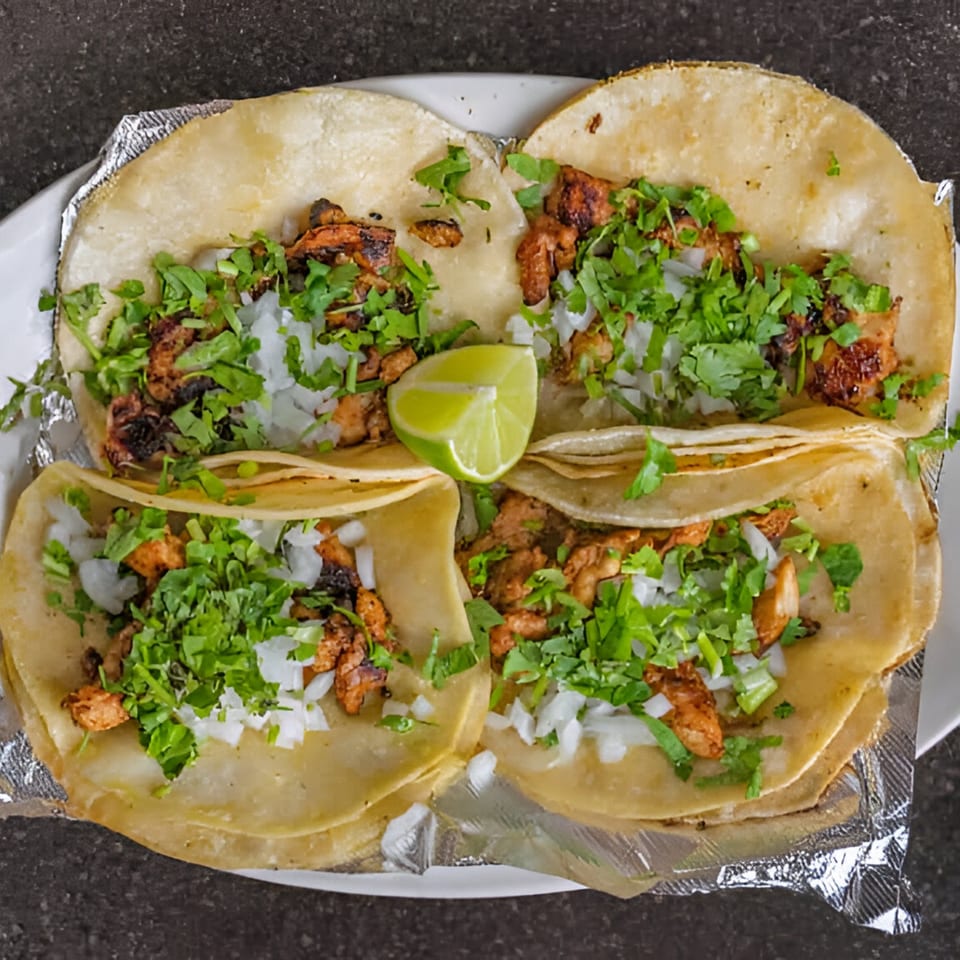 Four Guacamayas chicken tacos with cilantro, onions, and a lime wedge on a white plate.